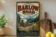 a vintage-style metal sign with a wagon traveling down a dirt road, surrounded by a mountainous landscape and pine trees.