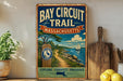 a vintage-style metal sign for the Bay Circuit Trail in Massachusetts, with a scenic illustration of a path leading to a sailboat on the water.