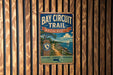 A vintage metal sign for the Bay Circuit Trail in Massachusetts, featuring a scenic image of a beach and a sailboat.