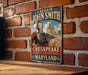 A vintage-style metal sign featuring a portrait of a bearded man in historical attire, with the text "CAPTAIN JOHN SMITH" and "CHESAPEAKE NATIONAL HISTORIC TRAIL MARYLAND" displayed on it.