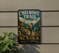 A vintage metal sign for the Chilkoot Trail Historic Corridor in Alaska, featuring a hiker on a trail surrounded by mountains and a scenic landscape.