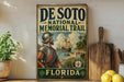 A vintage-style metal sign for the De Soto National Memorial Trail in Florida, featuring a painting of a man in historical attire and a depiction of a ship on the water.