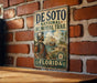 A vintage metal sign for the De Soto National Memorial Trail in Florida, featuring a historical illustration of a man in period attire and a ship.