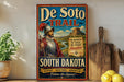 A vintage-style metal sign advertising the De Soto Trail in South Dakota, featuring a knight in armor and a mountainous landscape.