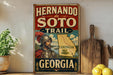 A vintage-style metal sign for the Hernando de Soto Trail in Georgia, featuring a map and a portrait of a man in historical attire.