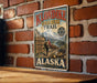 A vintage-style metal sign advertising Klondike Gold Rush Trail in Alaska, featuring a mountain landscape and a pan for gold.