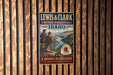 A vintage-style metal sign for the Lewis and Clark National Historic Trail in Idaho, featuring a scenic illustration of two hikers on a mountain trail.