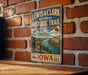 A vintage-style metal sign for the Lewis & Clark National Historic Trail, featuring a scenic illustration of a river and mountains, mounted on a brick wall.