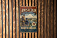 A vintage metal sign for the Lewis & Clark National Historic Trail, featuring a scenic illustration of a man and a woman standing on a cliff overlooking a river.