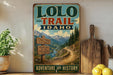 A vintage-style sign for the Lolo Trail in Idaho, featuring a scenic mountain landscape with a winding trail and a signpost.