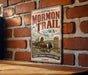 A vintage-style metal sign with the text "Historic Mormon Trail Iowa" and an illustration of a wagon train on a dirt road, mounted on a brick wall.
