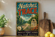 A vintage-style metal sign for Natchez Trace, Alabama, featuring a scenic road and landscape, is displayed on a shelf next to a wooden cutting board and some lemons.