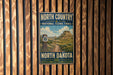 A vintage-style metal sign for the North Country National Scenic Trail in North Dakota, featuring a scenic landscape with mountains and a trail.