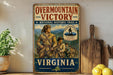 a vintage-style metal sign with the text "Overmountain Victory" and "National Historic Trail" on it, along with an illustration of a group of people in historical clothing. The sign is displayed on a shelf next to a wooden cutting board and a potted plant.