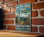 A vintage-style metal sign for the Pacific Crest Trail Historic Corridor in Washington, featuring a mountain landscape and a trail marker.