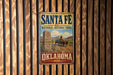 A vintage-style metal sign for Santa Fe National Historic Trail in Oklahoma, featuring a wagon and a monument, mounted on a wooden wall.