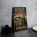 A vintage-style metal sign with the text "WASHINGTON-ROCCHAMBEAU REVOLUTIONARY ROUTE VIRGINIA" and an illustration of two men on horseback, one of whom is holding a flag. The sign is placed on a gray surface, with a stack of black camera lenses and a white dish visible in the background.