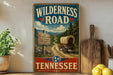 A vintage-style sign for the Wilderness Road in Tennessee, featuring a wagon on a dirt road surrounded by trees and mountains.
