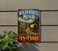 A vintage-style metal sign for the Wilderness Road in Tennessee, featuring a wagon on a dirt road surrounded by trees and mountains.