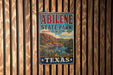 A vintage metal sign for Abilene State Park in Texas, featuring a scenic landscape with mountains, water, and desert vegetation.