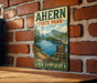A vintage metal sign for Ahern State Park in New Hampshire, featuring a scenic mountain landscape with a lake and pine trees.