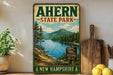 a vintage-style sign for Ahern State Park in New Hampshire, with a scenic landscape depicting a lake surrounded by pine trees and mountains.