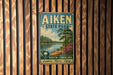 A vintage metal sign for Aiken State Park, featuring a scenic landscape with a lake, trees, and a person walking along the shore.