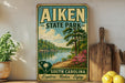 a vintage-style sign for Aiken State Park, with a scenic landscape depicting a lake, trees, and mountains. The sign is displayed on a shelf next to a wooden cutting board and some potted plants.