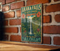 A vintage-style metal sign for Akaka Falls State Park in Hawaii, featuring a waterfall and tropical foliage, is displayed on a brick wall.