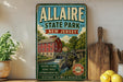 A vintage-style metal sign for Allaire State Park in New Jersey, featuring a red brick building, a watermill, and scenic trails, is displayed on a shelf next to a wooden cutting board and some lemons.
