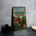 A vintage-style metal sign for Allaire State Park in New Jersey, featuring a red brick building and a watermill, is displayed on a gray countertop next to some camera equipment.