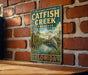 A vintage-style metal sign for Catfish Creek Preserve in Florida, featuring an illustration of a bird standing on a bridge overlooking a pond.