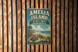 A vintage-style metal sign for Amelia Island State Park in Florida, featuring a beach scene with palm trees, a turtle, and a bird.