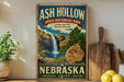 A vintage-style sign for Ash Hollow State Historical Park in Nebraska, featuring a waterfall and a rock formation, is displayed on a shelf next to a wooden cutting board and some lemons.