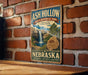 A vintage-style metal sign for Ash Hollow State Historical Park in Nebraska, featuring a waterfall and a buffalo, mounted on a brick wall.