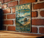 A vintage-style metal sign for Bandon State Natural Area in Oregon, featuring a scenic landscape with rocky cliffs, a beach, and a forest.