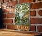 A vintage-style metal sign for Bastrop State Park in Texas, featuring a scenic illustration of a river and lush greenery.