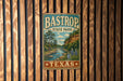 A vintage metal sign for Bastrop State Park in Texas, featuring a scenic landscape with a river and mountains.