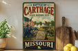 A vintage-style metal sign depicting a battle scene in Missouri, with soldiers and a cannon, is displayed on a shelf next to a wooden cutting board and some lemons.