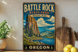A vintage-style metal sign for Battle Rock State Park in Oregon, featuring a scenic mountain landscape with a river and pine trees.