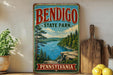 A vintage-style metal sign for Bendigo State Park in Pennsylvania, featuring a scenic landscape with a river, pine trees, and a rocky cliff.