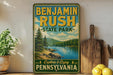 A vintage-style sign for Benjamin Rush State Park in Pennsylvania, featuring a scenic landscape with pine trees, a lake, and mountains.