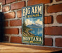 A vintage-style metal sign for Big Arm State Park in Montana, featuring a scenic landscape with mountains, a lake, and a sailboat.