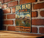 A vintage-style metal sign for Big Bend Ranch, Texas, featuring a desert landscape with mountains, cacti, and a river.