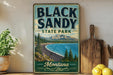 A vintage-style sign for Black Sandy State Park in Montana, featuring a scenic landscape with mountains, pine trees, and a lake.