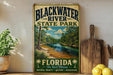 A vintage-style sign for Blackwater River State Park in Florida, featuring a scenic image of the river surrounded by lush greenery.