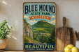 A vintage-style sign for Blue Mound State Park in Wisconsin, featuring a scenic mountain landscape with a small village in the distance.