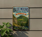 A vintage-style metal sign for Blue Mound State Park in Wisconsin, featuring a scenic mountain landscape with trees and a winding path.