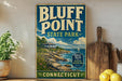 A vintage-style sign for Bluff Point State Park in Connecticut, featuring a coastal landscape with a house, boats, and a lighthouse.