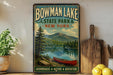 A vintage-style sign for Bowman Lake State Park in New York, featuring a scenic image of a lake, pine trees, and a canoe.